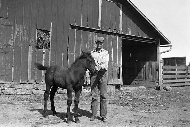 A black-and-white photo of a man in a flat cap holding a young foal in front of a wooden barn